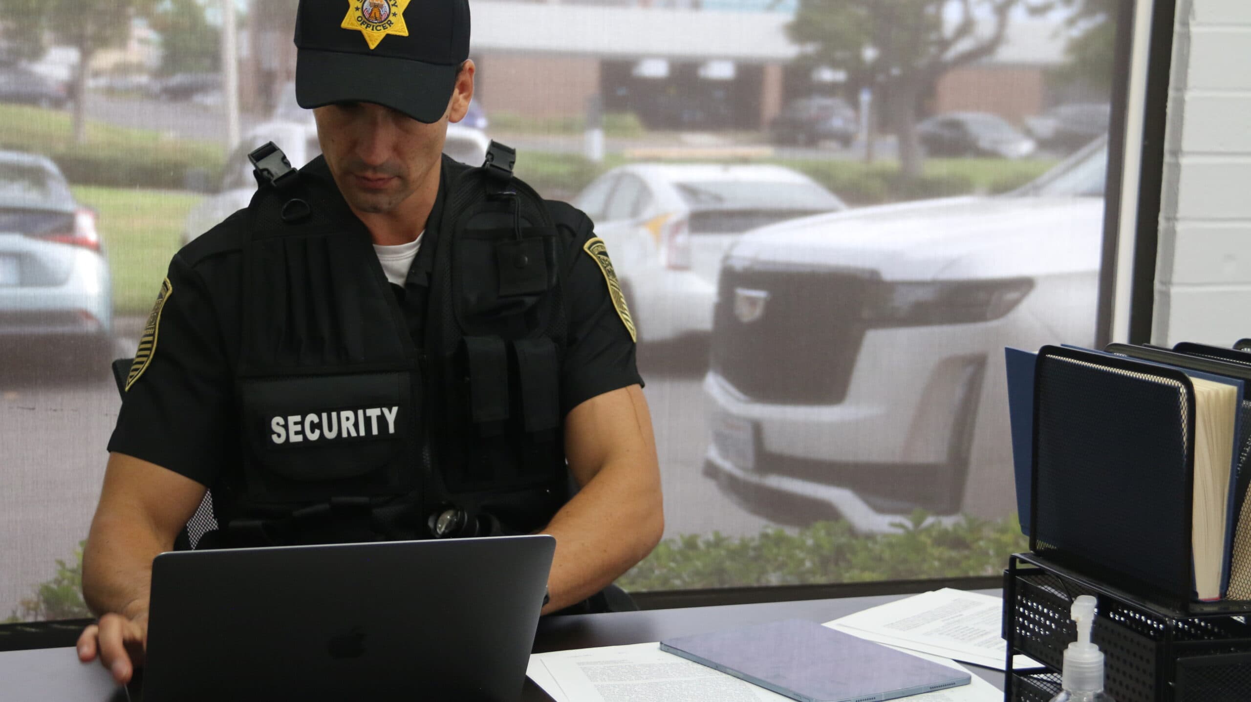A uniformed hospital security guard stands attentively near a hospital entrance, monitoring access while assisting an elderly patient in a wheelchair. In the background, clear signage directs visitors to emergency services, while a wall-mounted security camera reinforces the facility's safety measures. The guard's radio and professional demeanor convey vigilance, while his helpful posture demonstrates the dual role of protection and patient support in healthcare environments