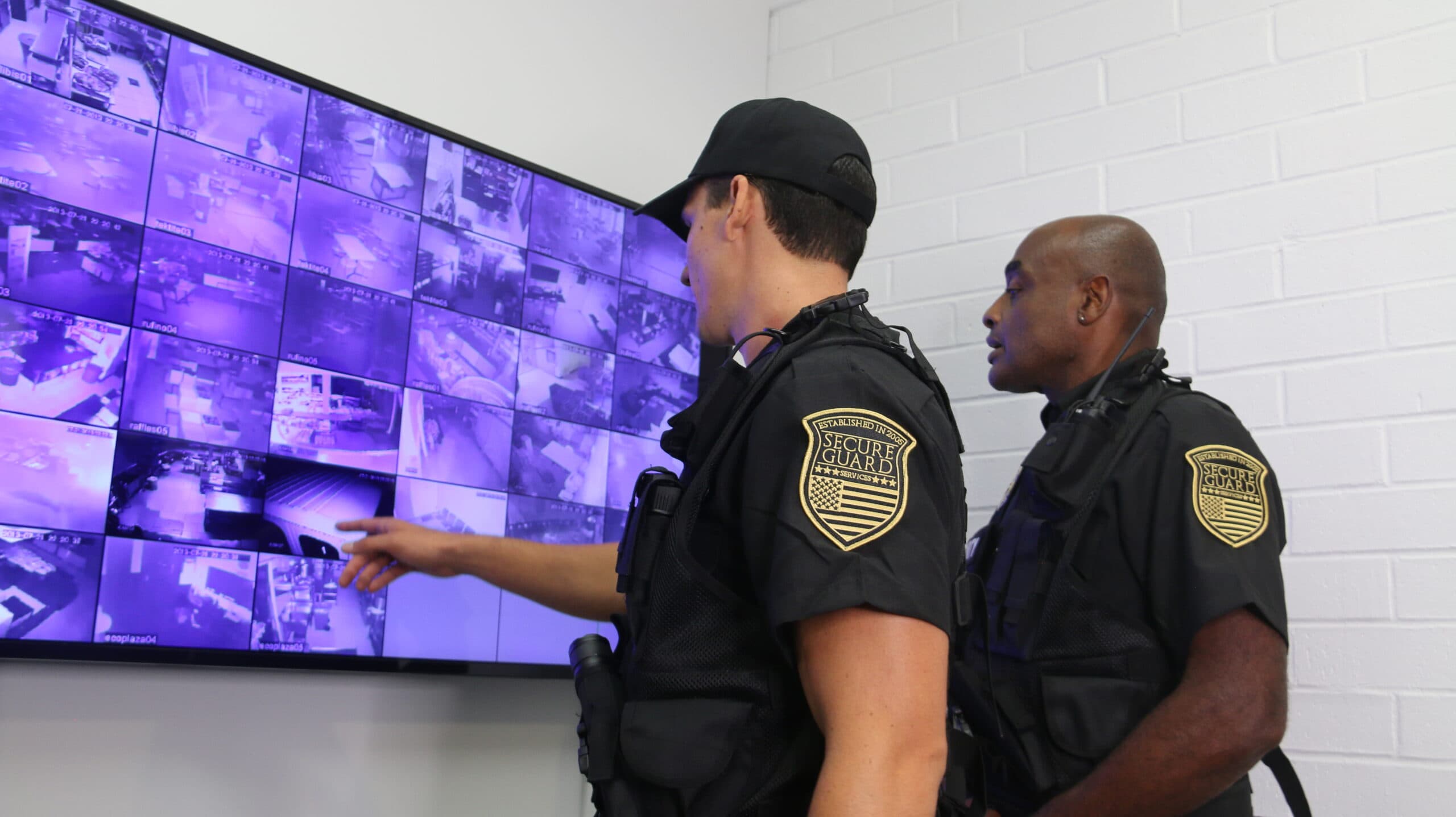 Two uniformed security officers monitor multiple live surveillance camera feeds on a large screen, ensuring property safety and real-time threat detection.