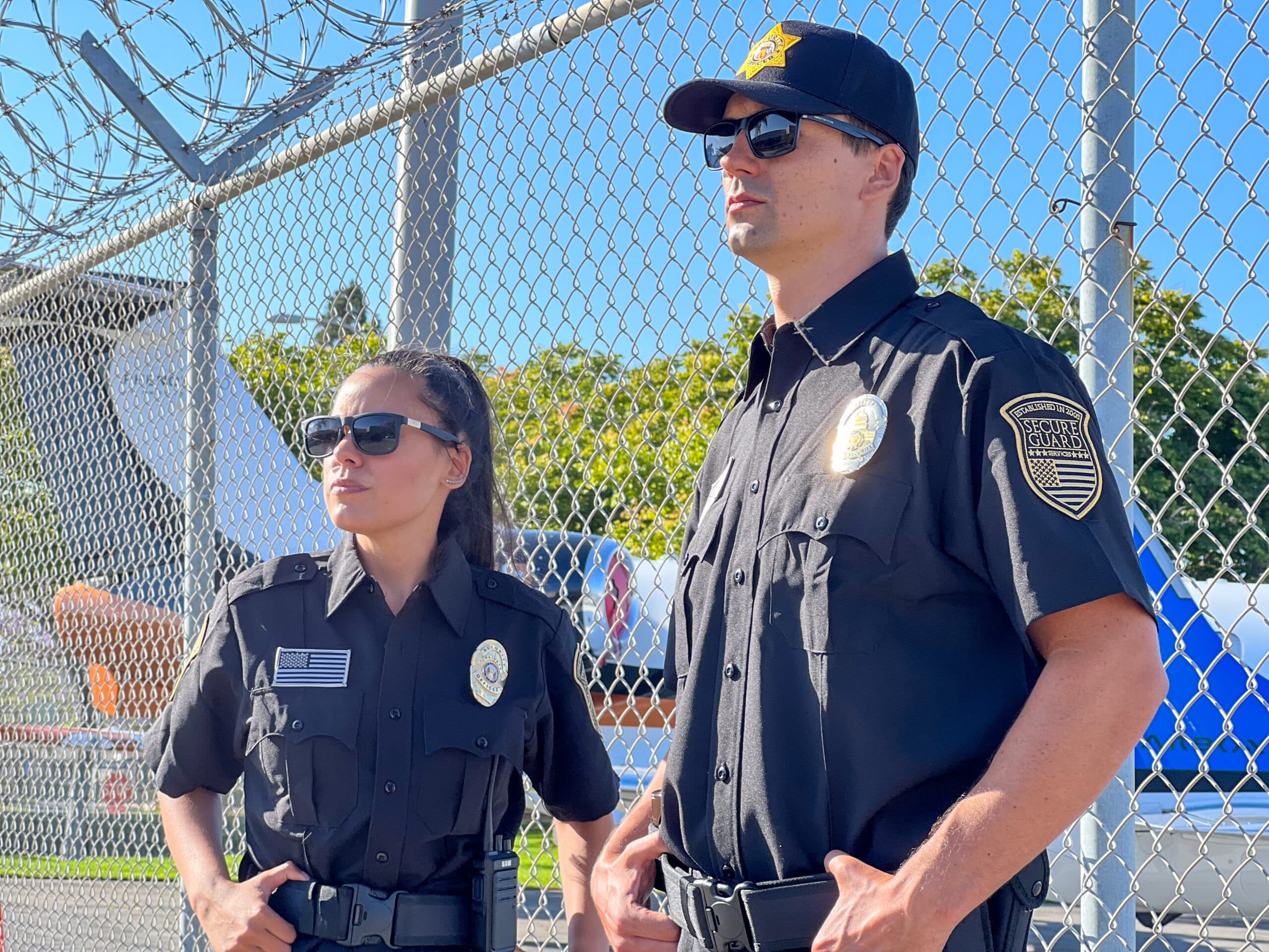 Two professional security guards standing alert near a secured fence, representing quality security services and what to look for when hiring a security company